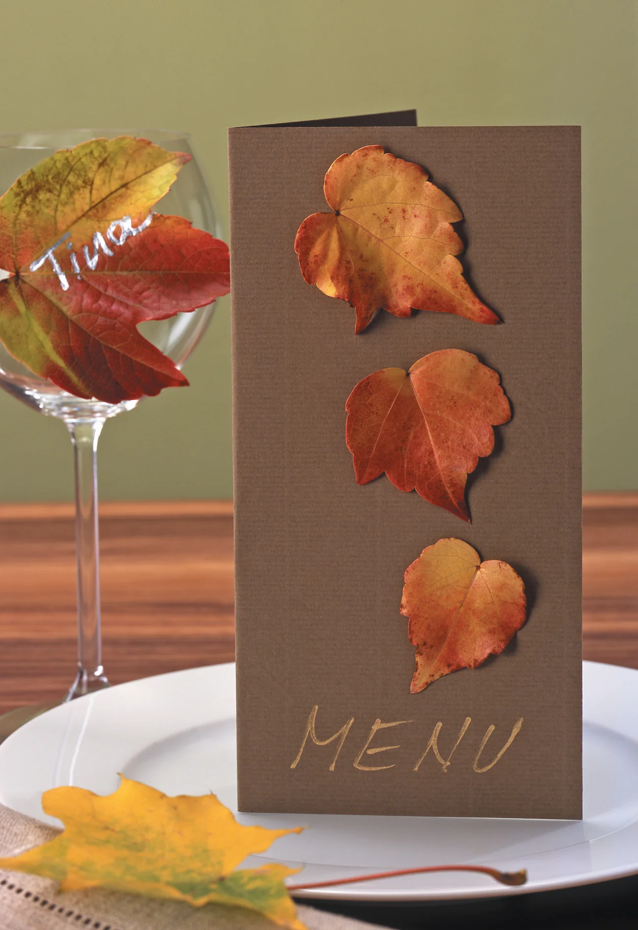 Een bruine menukaart met MENU onderaan, toont drie herfstbladeren, allemaal vastgemaakt met tesa tape. Het is op een wit bord gelegd. Een wijnglas bevat een rood blad met de naam Tina vastgemaakt met tesa tape. Een geel blad is met tesa tape aan de tafel bevestigd. De achtergrond is olijfgroen. (This text has been generated by AI)