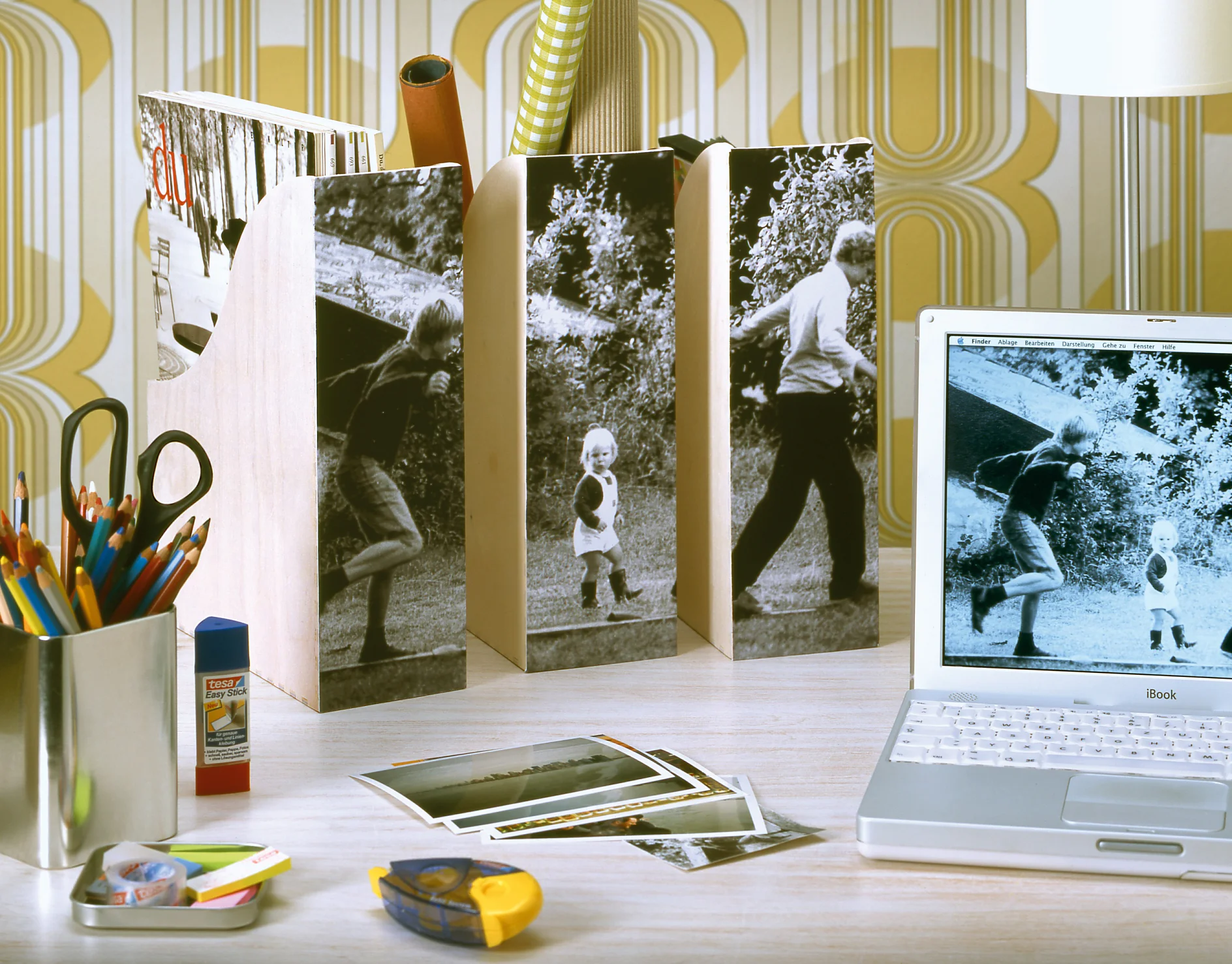 Een rommelig bureau met een laptop waarop een zwart-witfoto staat. Ernaast staan houten tijdschriftenhouders met zwart-witafbeeldingen. Tot de spullen behoren kleurpotloden, een lijmstift, tesa-tape, fotos en decoratief behang op de achtergrond. (This text has been generated by AI)