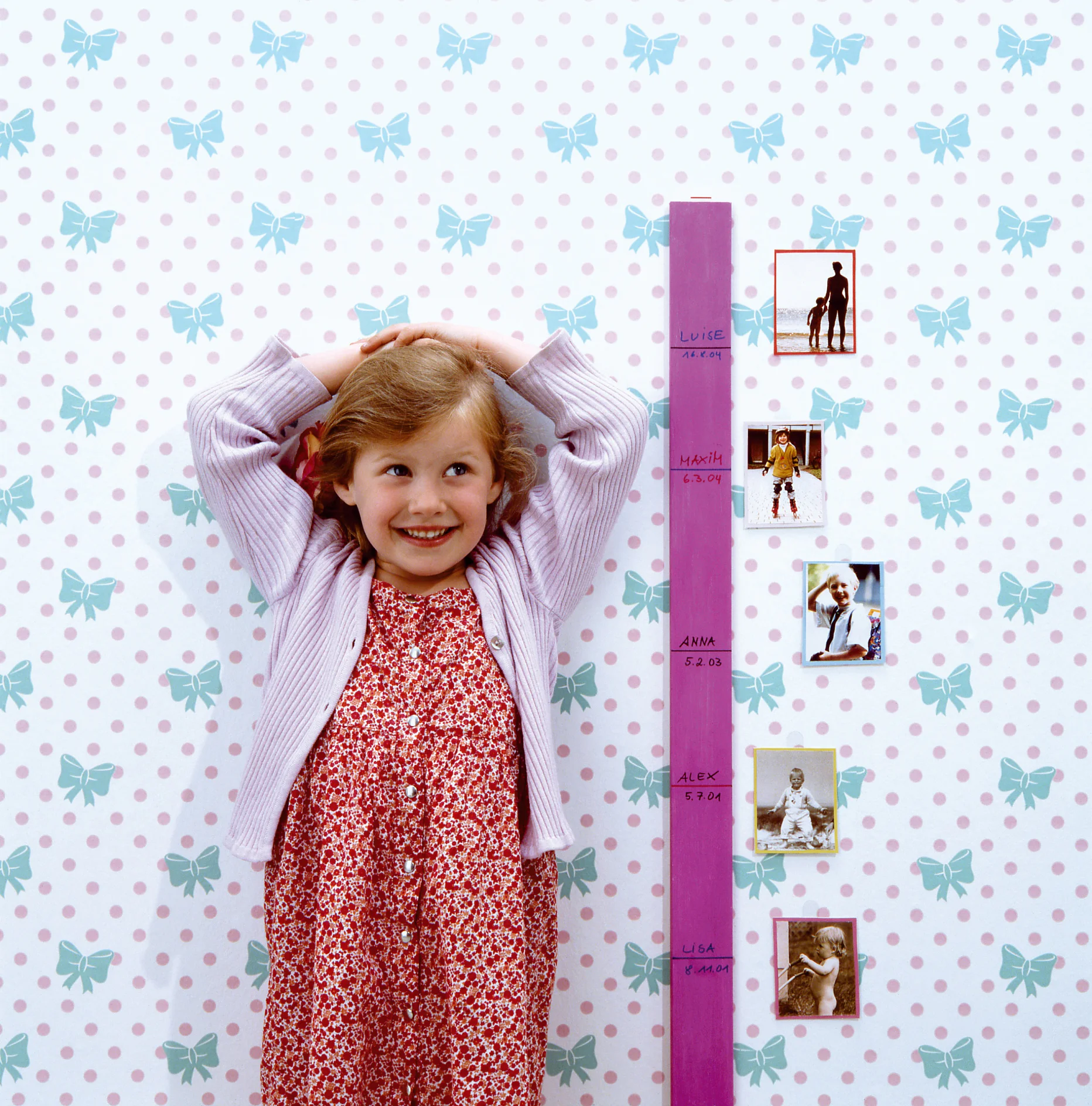 Een jong meisje in een rode jurk en een gestreepte trui lacht terwijl ze naast een groeimeter op een muur staat met een strik- en stippenpatroon. De meter, bij elkaar gehouden met tesa-tape, bevat familiefotos en namen die verschillende lengtes aangeven. (This text has been generated by AI)