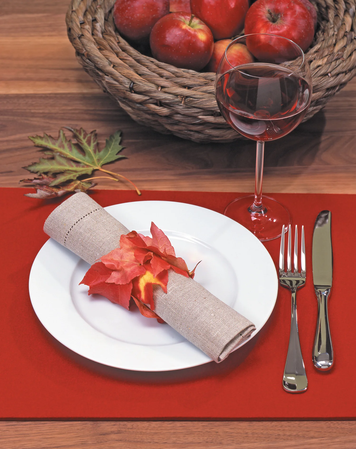 Een eetopstelling met een wit bord op een rode placemat, met een beige servet omwikkeld met rode bladeren en vastgezet met tesa tape. Rechts liggen een vork en mes. Een glas rode wijn staat erboven, met een gevlochten mand gevuld met rode appels op de achtergrond. (This text has been generated by AI)