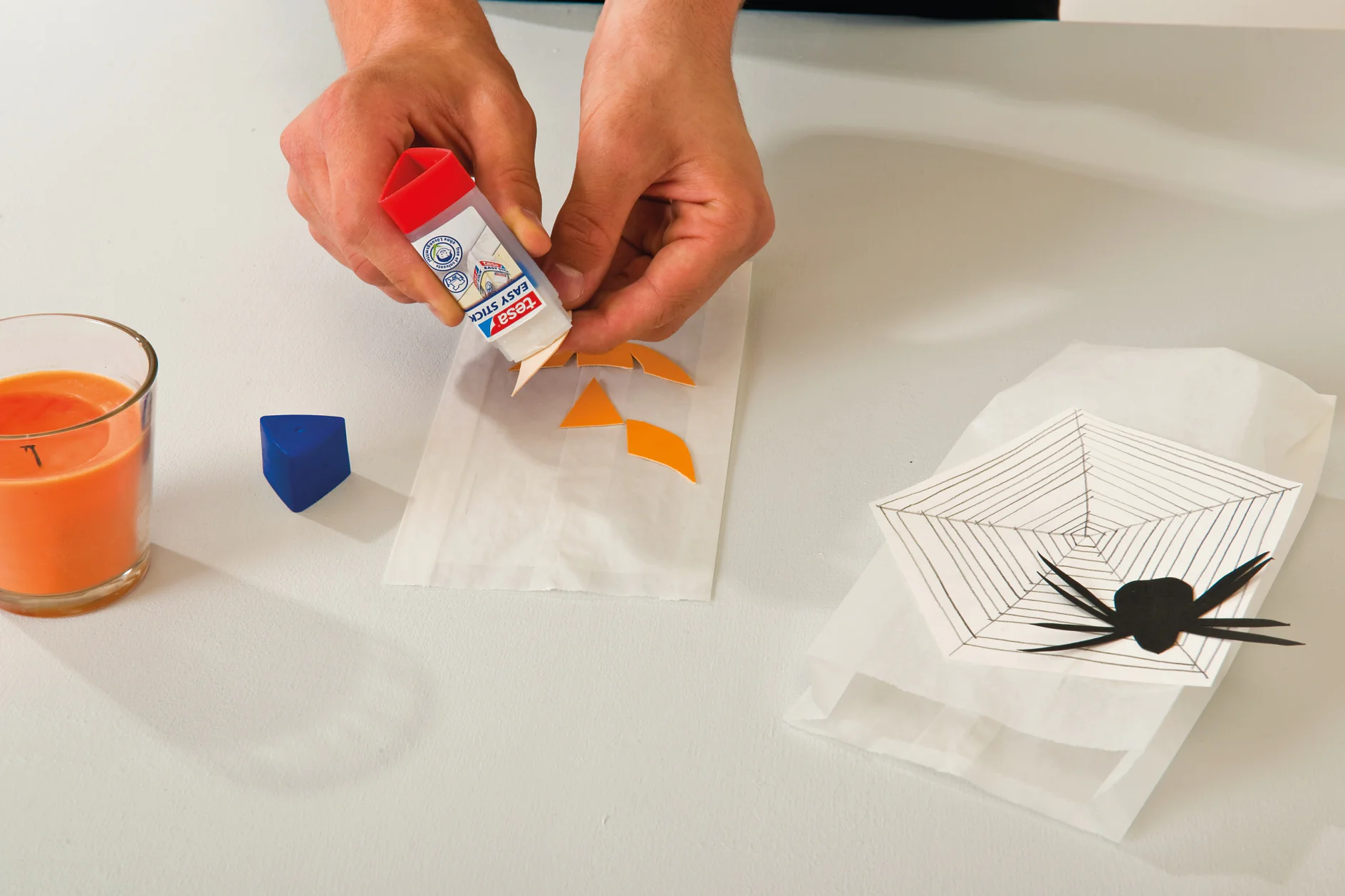Handen plakken oranje papieren vormen op een wit oppervlak naast een kaars, een blauwe dop en een spinnenwebontwerp met een zwarte spin-uitsparing. Een rode en witte tesa-tape-stick wordt gebruikt. (This text has been generated by AI)