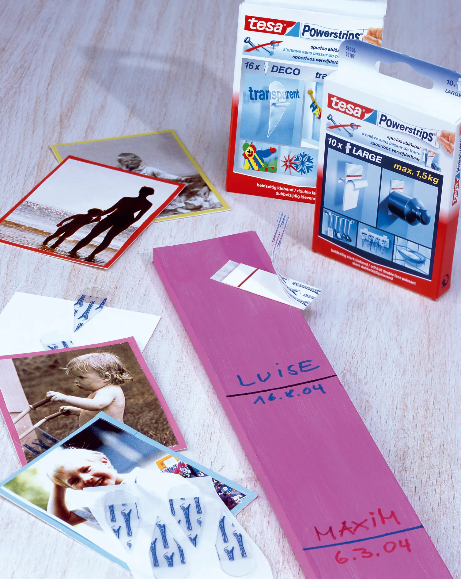 Op een tafel liggen een verzameling persoonlijke fotos, een roze houten blok met het label Luise 16.6.04 en een blauw blok met het label Maxim 6.3.04. Ernaast liggen verpakkingen van tesa Powerstrips, met verschillende verpakkingsontwerpen en details. (This text has been generated by AI)