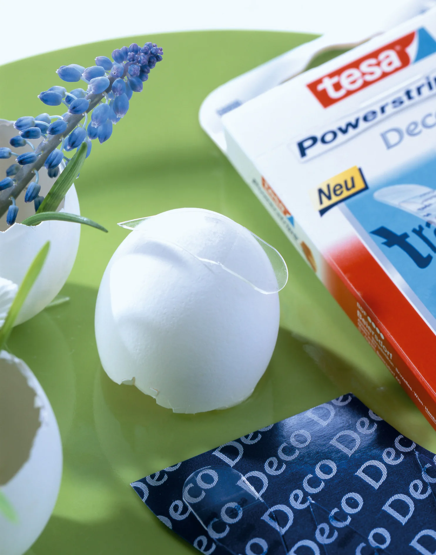 Een close-up van een gebarsten witte eierschaal op een groen oppervlak, met een kleine paarse bloem in de buurt. Een doos tesa Powerstrips Deco is zichtbaar op de achtergrond, gedeeltelijk geopend, met een decoratief stuk stof met het label Deco. (This text has been generated by AI)