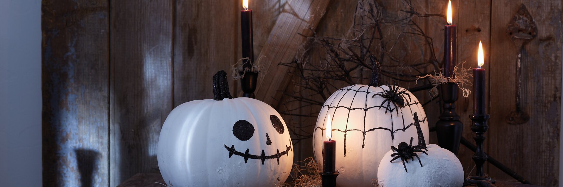 Een Halloween-tafereel met twee wit geschilderde pompoenen op een houten tafel. De pompoenen hebben zwarte jack-o-lanterngezichten. Er staan brandende zwarte kaarsen en verspreide takjes omheen. Er liggen een paar kleine boeken op tafel, kunstig vastgezet met wat onzichtbare tesa-tape om de opstelling stabieler te maken. Houten achtergrond met schaduwen, wat een sfeervolle setting creëert die perfect is voor de kerstsfeer. (This text has been generated by AI)
