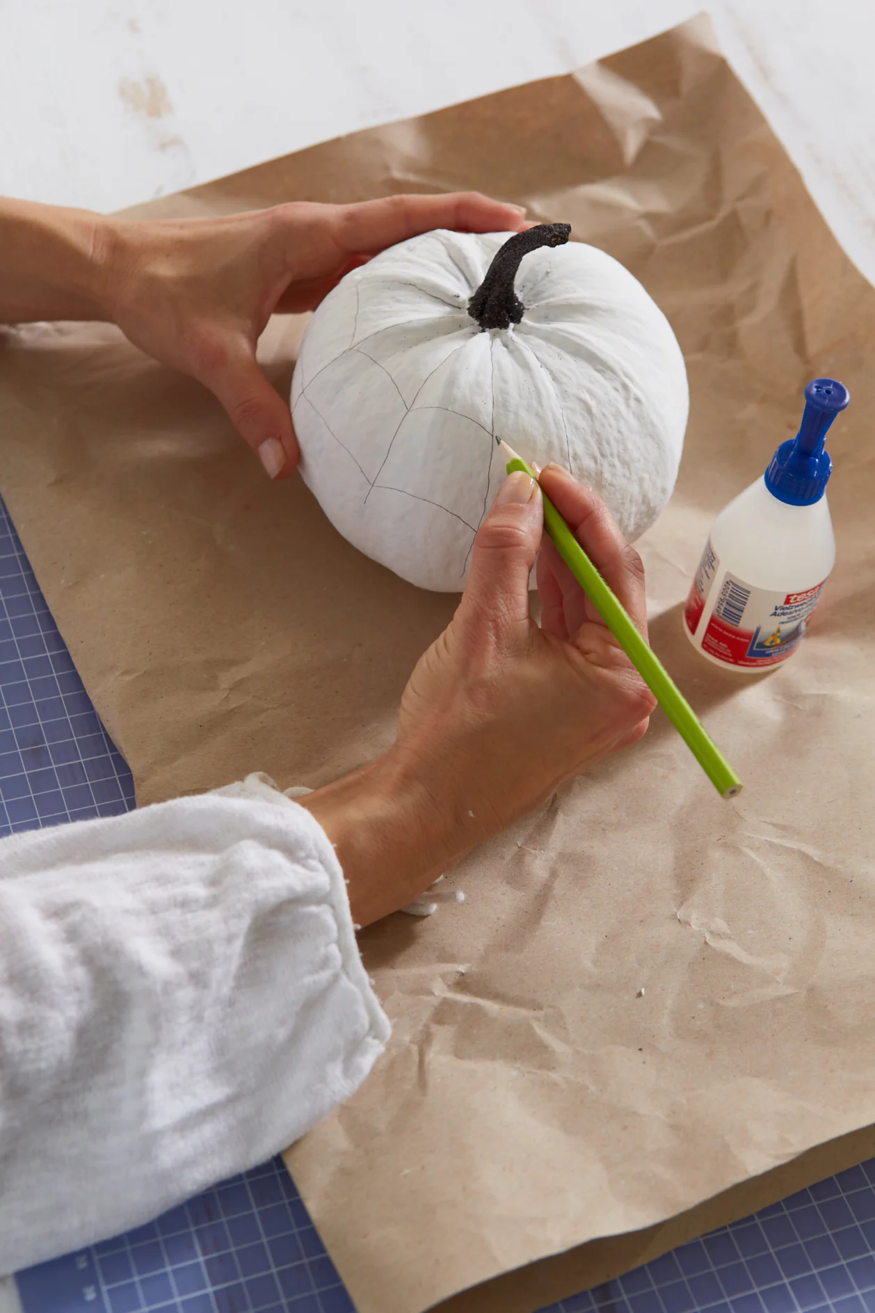 Persoon tekent een geometrisch patroon op een wit geschilderde pompoen met een groen potlood. De pompoen staat op bruin papier, naast een fles lijm en tesa tape. De persoon draagt een wit shirt met lange mouwen. (This text has been generated by AI)