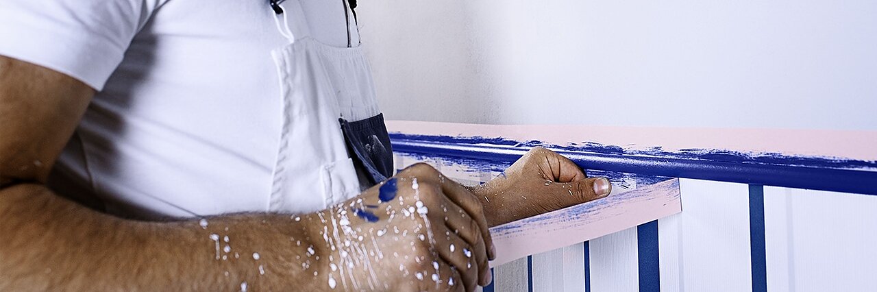 Een persoon in een witte overall schildert een muur met blauwe verf, met roze tesa tape voor een nette rand. Hun handen en overall hebben verfspatten. Ze brengen de verf aan op een witte muur met gelijkmatig verdeelde verticale strepen. (This text has been generated by AI)