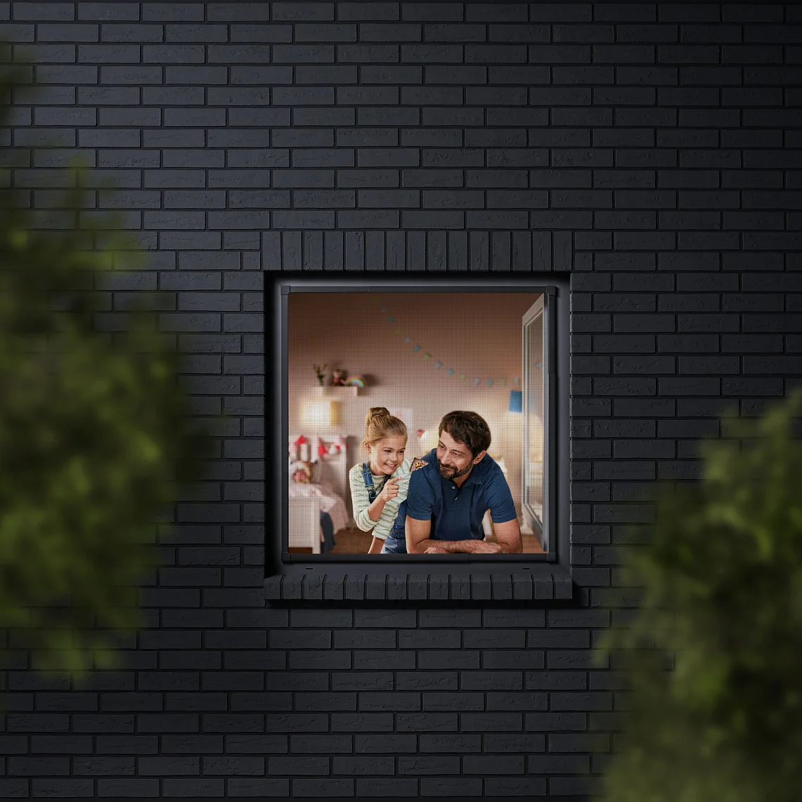 A man and a child are seen through a window, looking into a room decorated with colorful bunting. The building has dark brick walls, and the view is partially framed by blurred greenery. The man is wearing a blue shirt, and the child is sitting beside him. (This text has been generated by AI)
