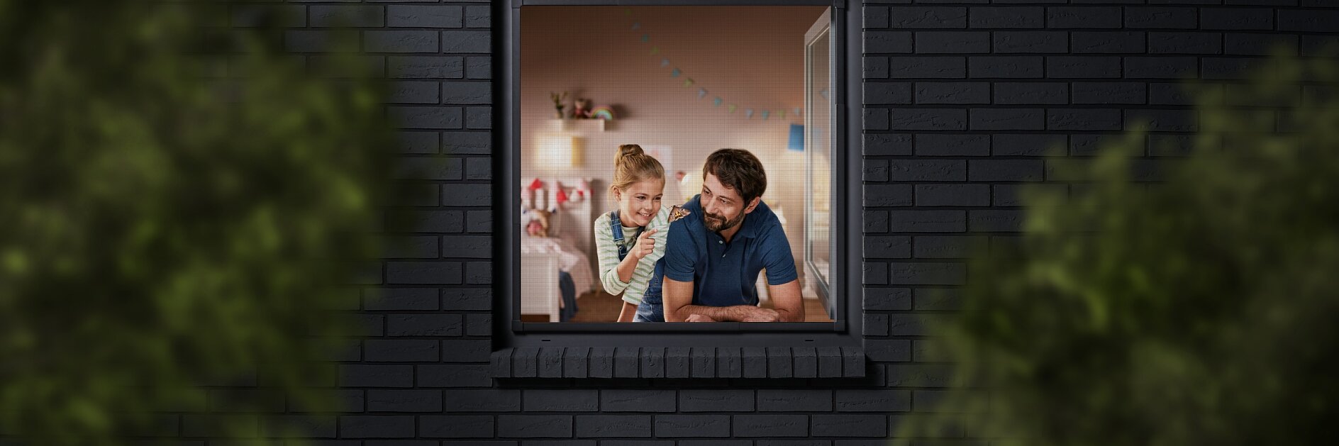 A man and a child are seen through a window, looking into a room decorated with colorful bunting. The building has dark brick walls, and the view is partially framed by blurred greenery. The man is wearing a blue shirt, and the child is sitting beside him. (This text has been generated by AI)