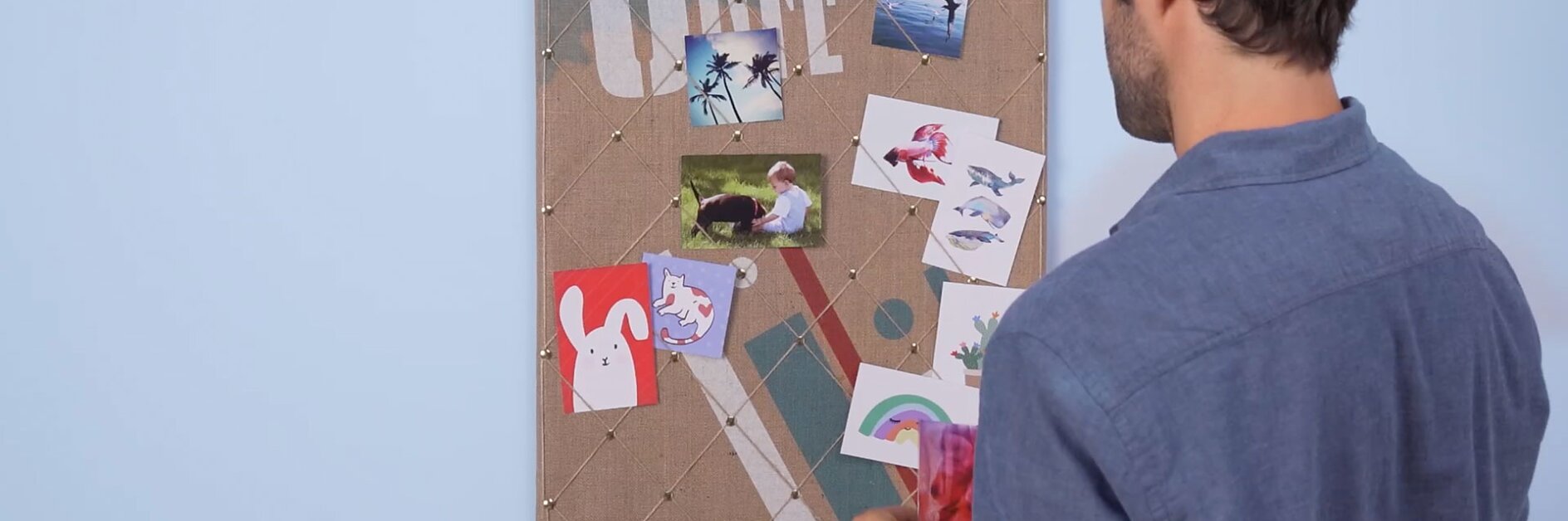 A person in a blue shirt stands facing a corkboard filled with various images and notes pinned to it using tesa tape. The board is against a light blue wall. The person is holding something red in their hand. (This text has been generated by AI)