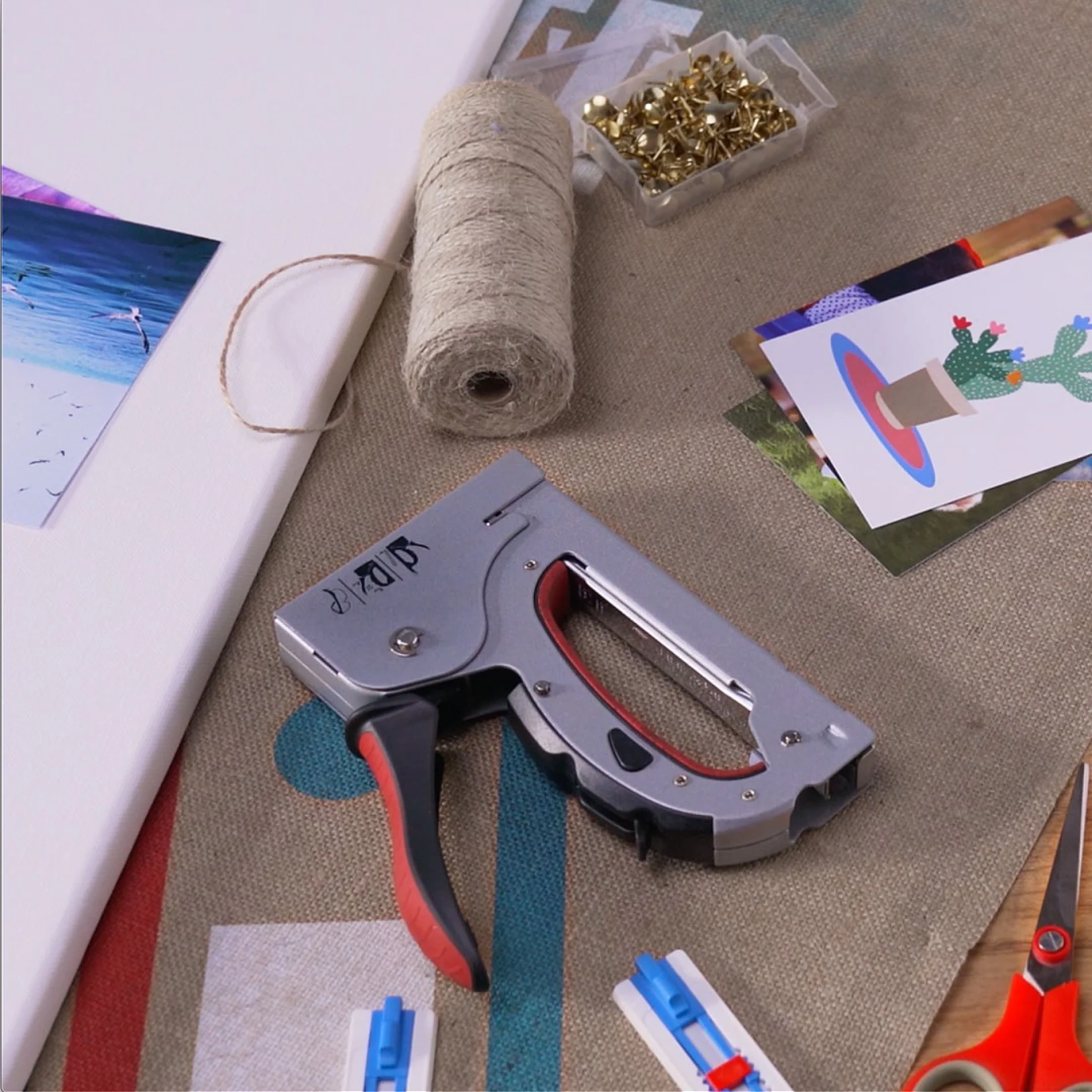 A staple gun is placed on a surface with a roll of twine beside it. Nearby are some photographs, push pins in a small container, and two blue-and-white plastic tools. Alongside these items, theres a roll of tesa tape. The background is a textured fabric. (This text has been generated by AI)
