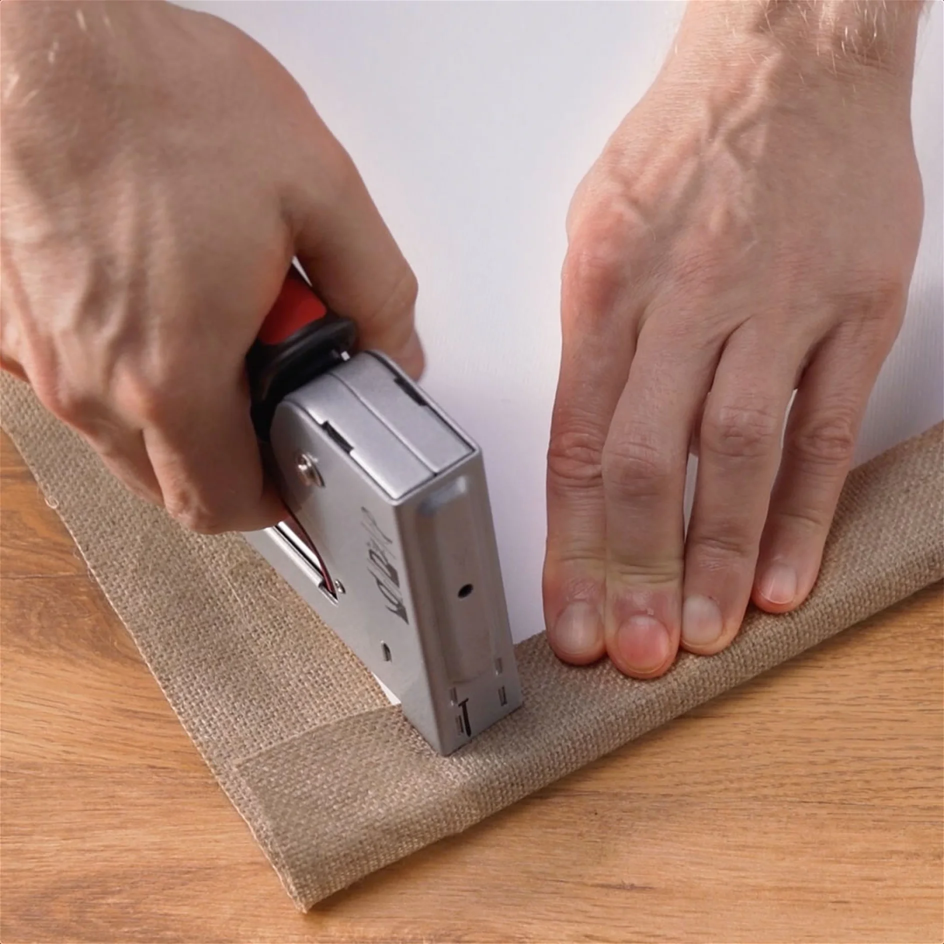 Hands are using tesa tape to secure burlap fabric onto a wooden surface. The right hand holds the fabric in place, while the left hand operates the tesa tape, pressing it down near the edge of the fabric. (This text has been generated by AI)