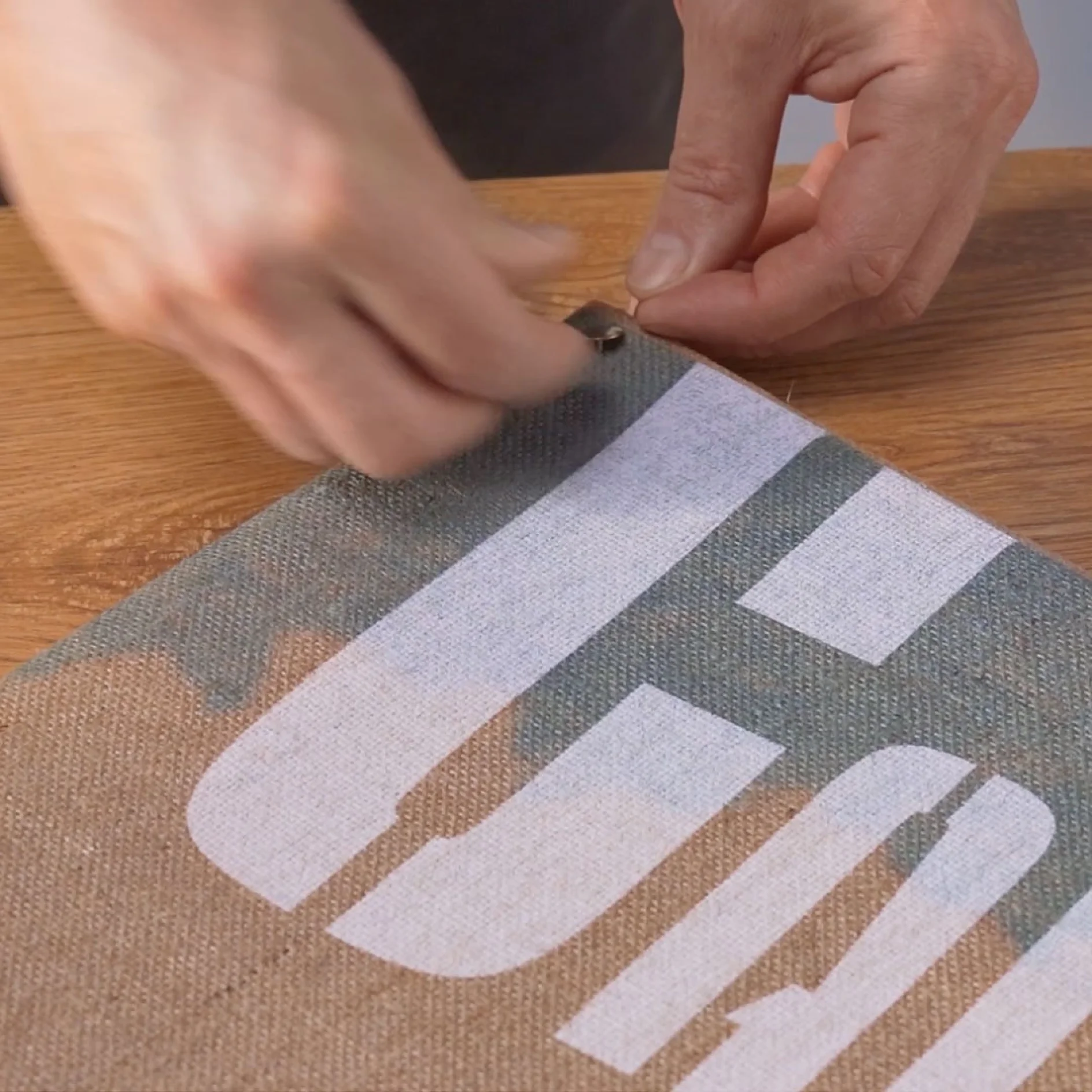 Hands are seen peeling off a protective film from a piece of fabric with large block letters printed on it using tesa tape. The fabric is on a wooden surface. (This text has been generated by AI)