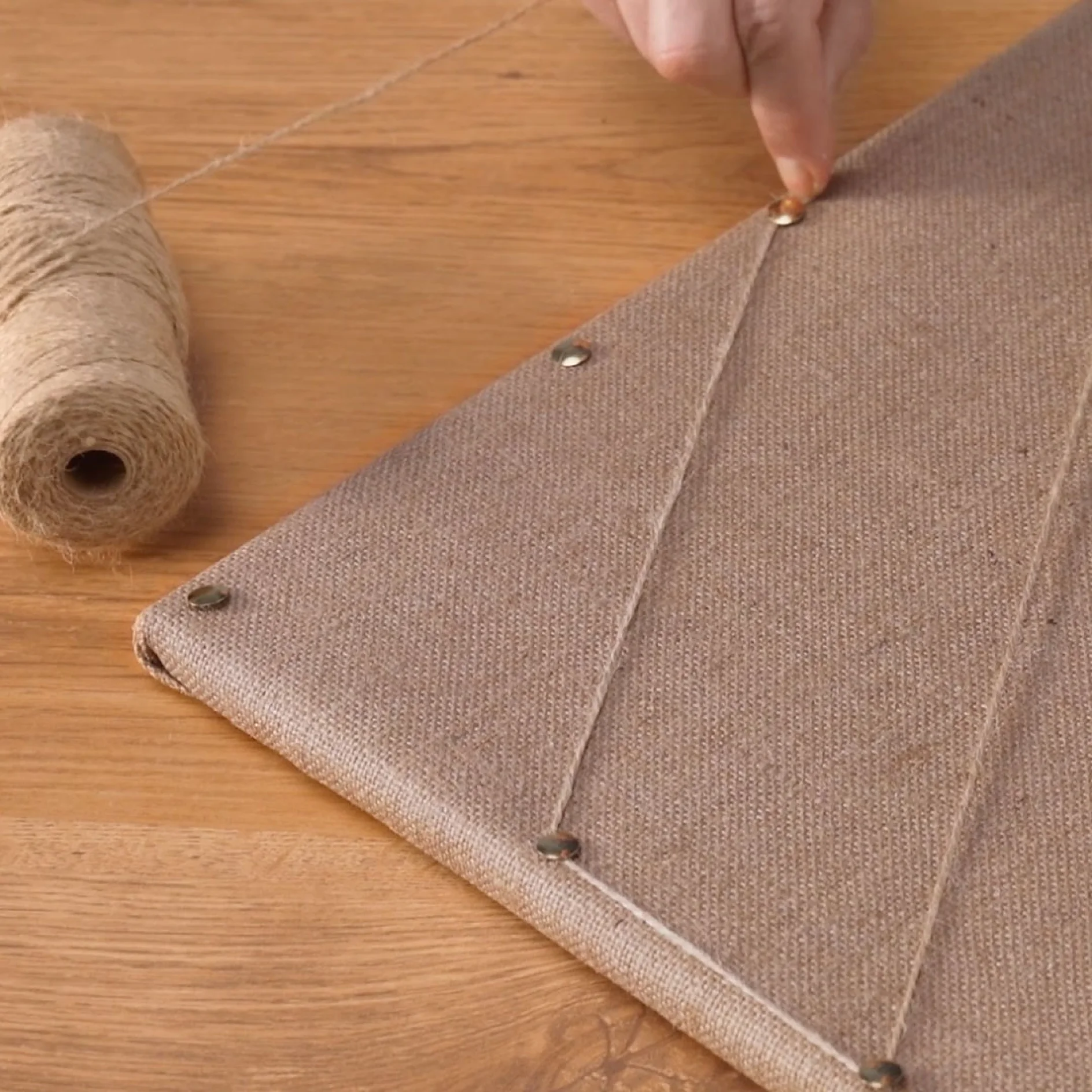 A persons hand is adjusting a piece of twine wrapped around a burlap-covered board. The board has metal pins securing the twine in a zigzag pattern. A spool of tesa tape is placed on the left on a wooden surface. (This text has been generated by AI)