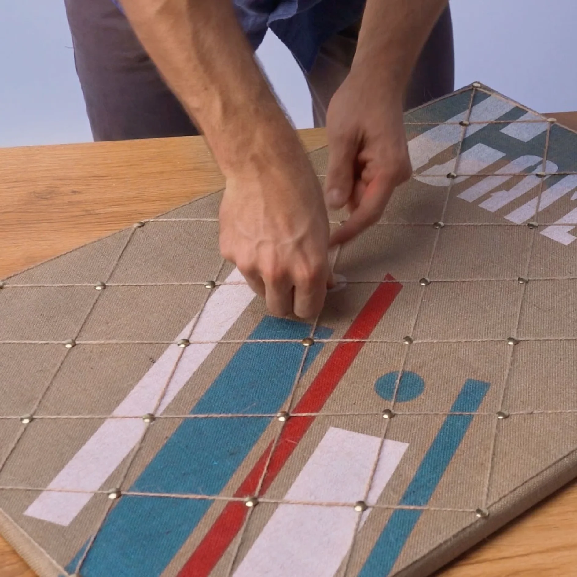 Two hands are working on a fabric-covered board with a geometric design. The board is secured to a wooden surface with tesa tape, creating a grid of twine and small nails, with red, blue, and white shapes visible on the fabric. (This text has been generated by AI)