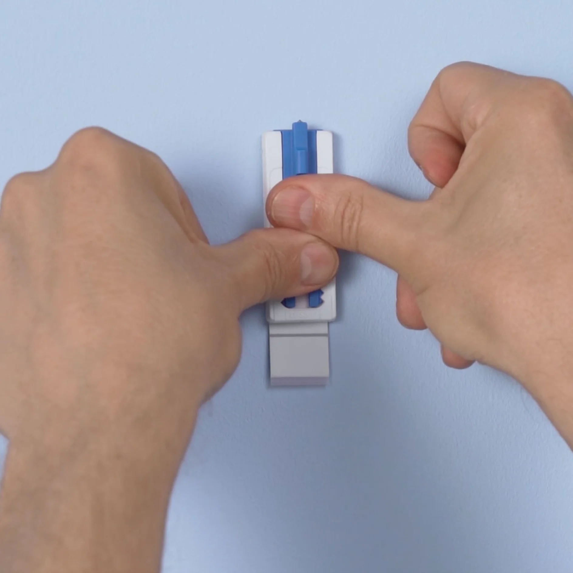 Two hands are seen securing a white wall hook with a blue locking mechanism onto a light blue wall. The person is pressing down on the hook, ensuring it is firmly attached. The hook appears to be using removable adhesive tesa tape. (This text has been generated by AI)