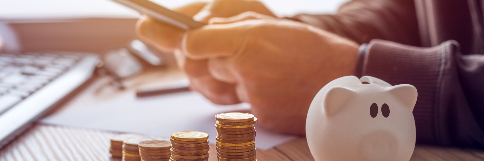 A person uses a smartphone at a desk with neatly stacked coins and a white piggy bank. The scene suggests financial planning or online banking, with a blurred keyboard and papers visible in the background.