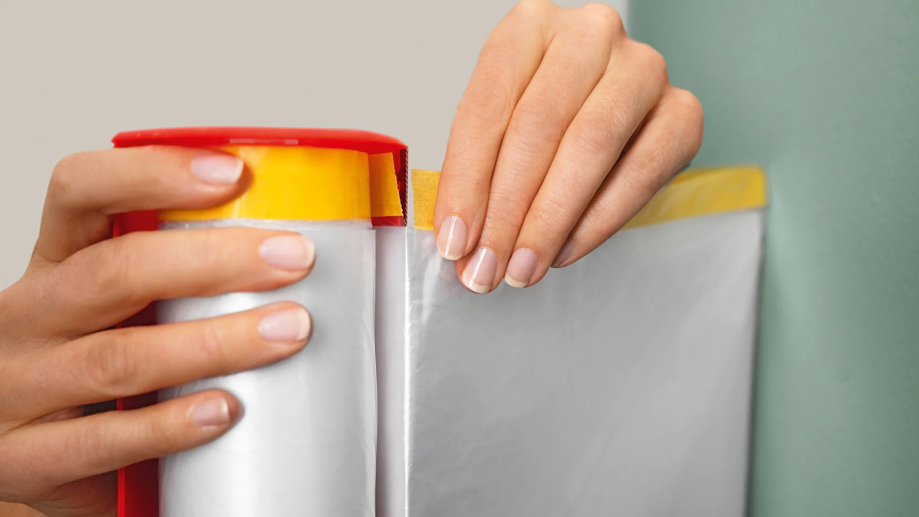 Handen trekken een vel doorzichtige plasticfolie uit een rood-gele tesa tape dispenser. De plasticfolie wordt uitgerold en de tesa tape dispenser wordt dicht bij een groene muur gehouden. (This text has been generated by AI)
