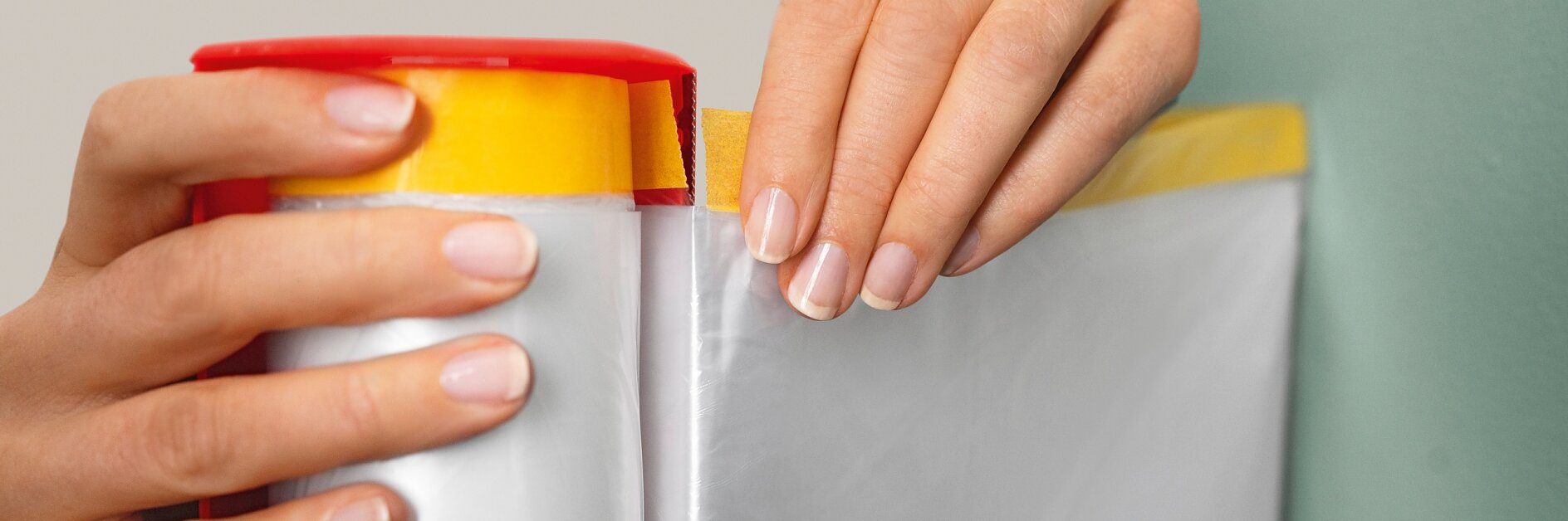 Handen trekken een vel doorzichtige plasticfolie uit een rood-gele tesa tape dispenser. De plasticfolie wordt uitgerold en de tesa tape dispenser wordt dicht bij een groene muur gehouden. (This text has been generated by AI)