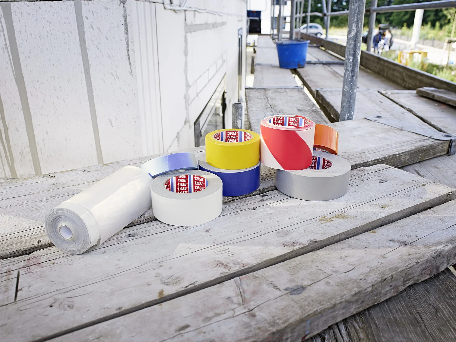 Several rolls of tesa tape in various colors, including white, yellow, blue, red, and orange, are placed on a wooden scaffold platform next to a roll of white material. Construction materials surround the area. (This text has been generated by AI)