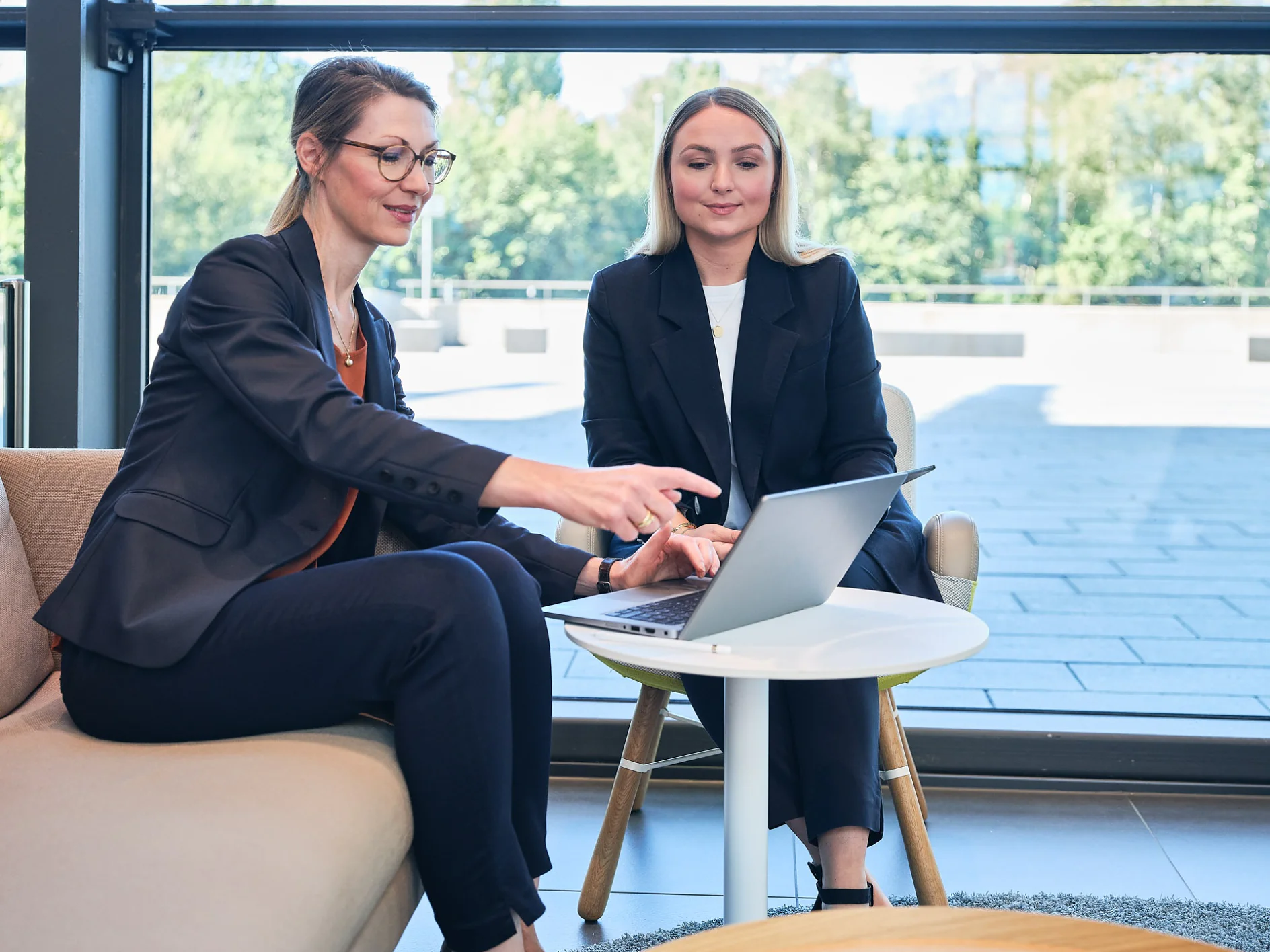 Two women in business attire are seated indoors at a small round table with a laptop. One woman is pointing at the screen while the other observes. Large windows in the background reveal a view of trees. (This text has been generated by AI)