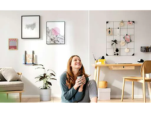 Een persoon met lang haar zit lachend op de grond in een lichte kamer, met een mok in de hand. Achter hen staat een bureau met tesa-tape en briefpapier, samen met een stoel en een prikbord versierd met items die met tesa-tape zijn bevestigd. Kunst en een plant versieren de ruimte. (This text has been generated by AI)