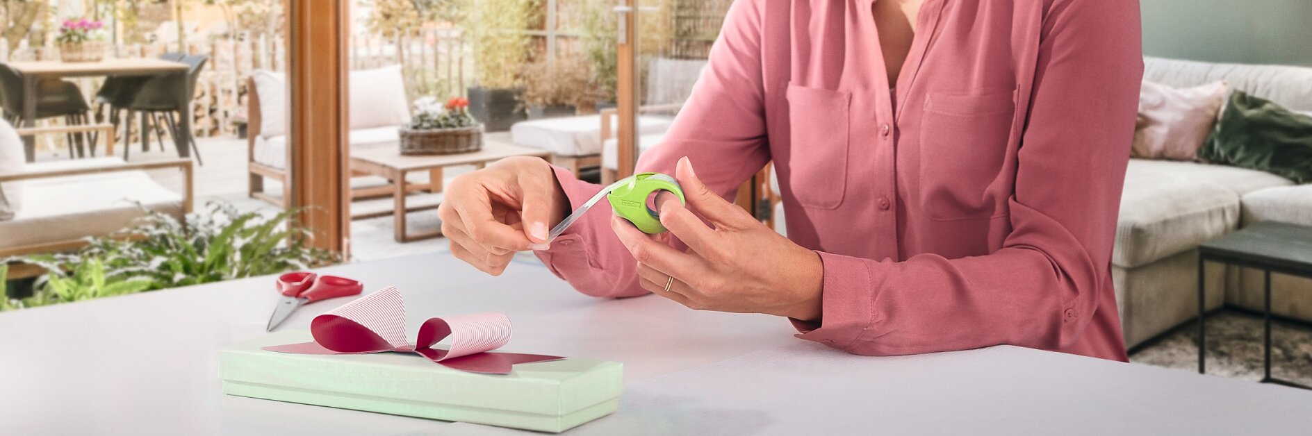 Een vrouw in een roze blouse zit aan een tafel in een lichte kamer met grote ramen, en wikkelt een smalle doos in met lichtgroen papier en een roze lint. Scharen en tesa-tape liggen op tafel. De achtergrond toont een gezellige woonkamer en patio. (This text has been generated by AI)