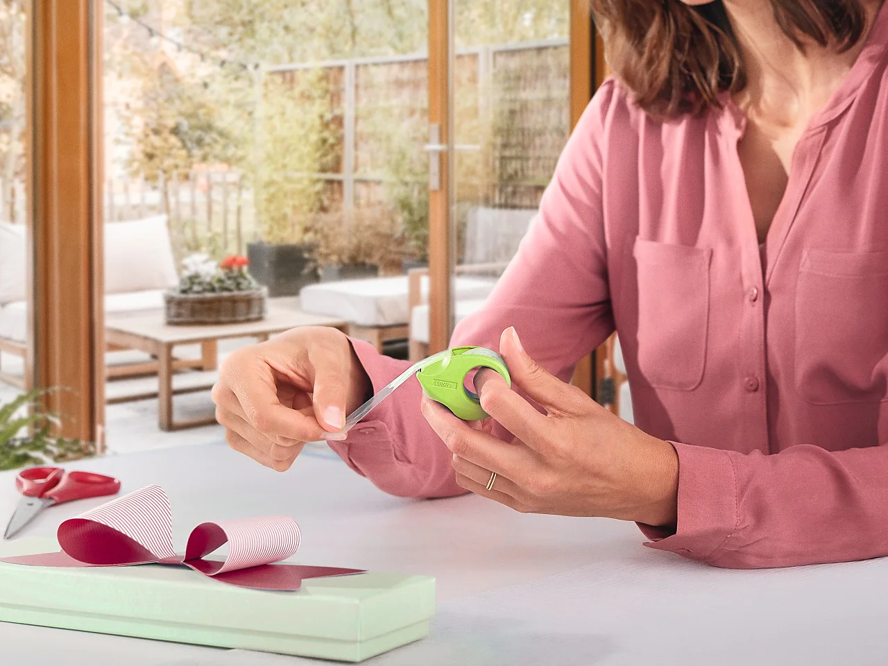 Een vrouw in een roze blouse zit aan een tafel in een lichte kamer met grote ramen, en wikkelt een smalle doos in met lichtgroen papier en een roze lint. Scharen en tesa-tape liggen op tafel. De achtergrond toont een gezellige woonkamer en patio. (This text has been generated by AI)