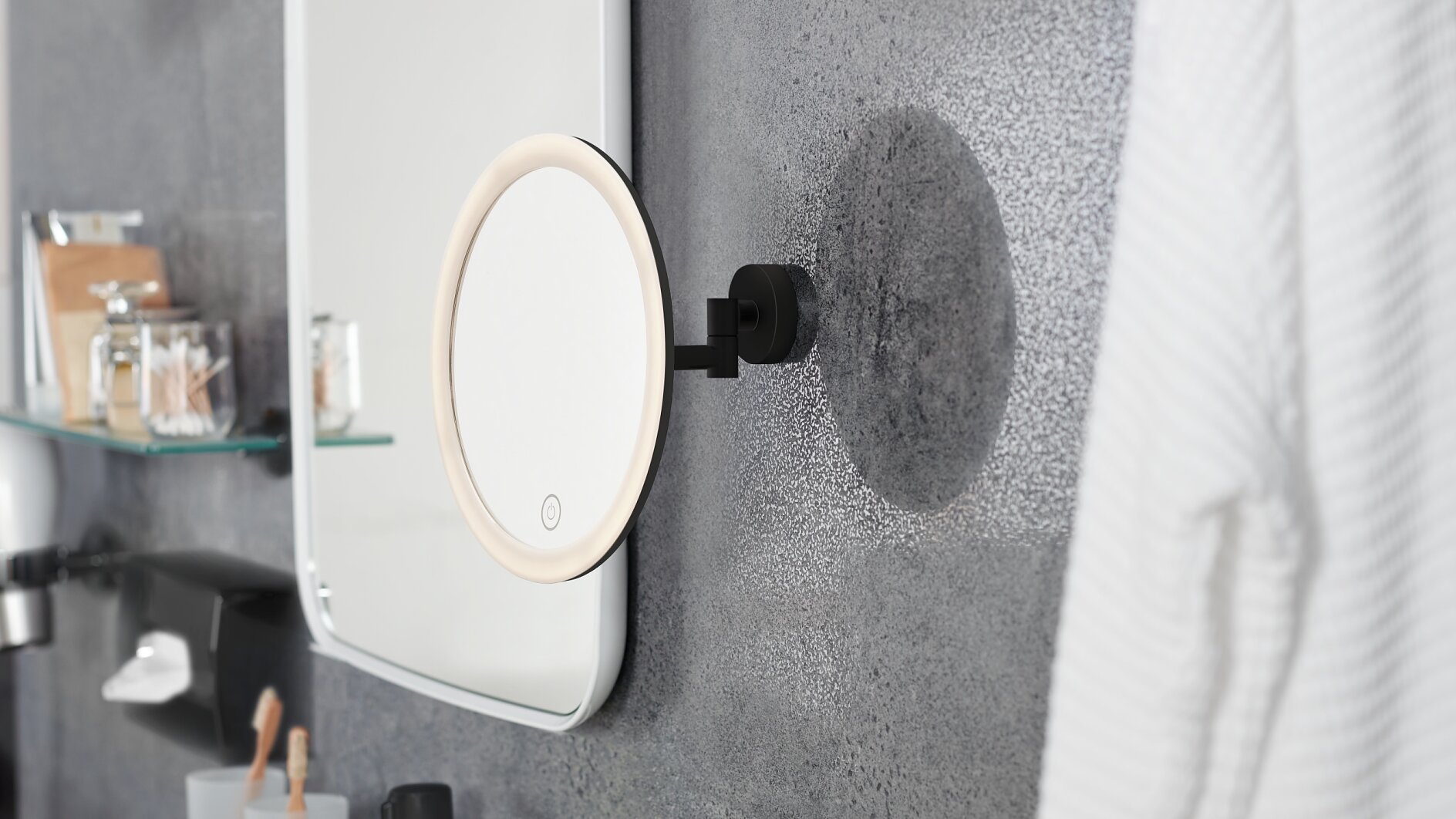Op een badkamermuur hangt een ronde, verlichte spiegel aan een verstelbare arm. Op de planken ernaast staan potten en toiletartikelen die zijn vastgezet met tesa tape. Rechts hangt een witte badjas. De muur heeft een gestructureerde, gespikkelde afwerking. (This text has been generated by AI)