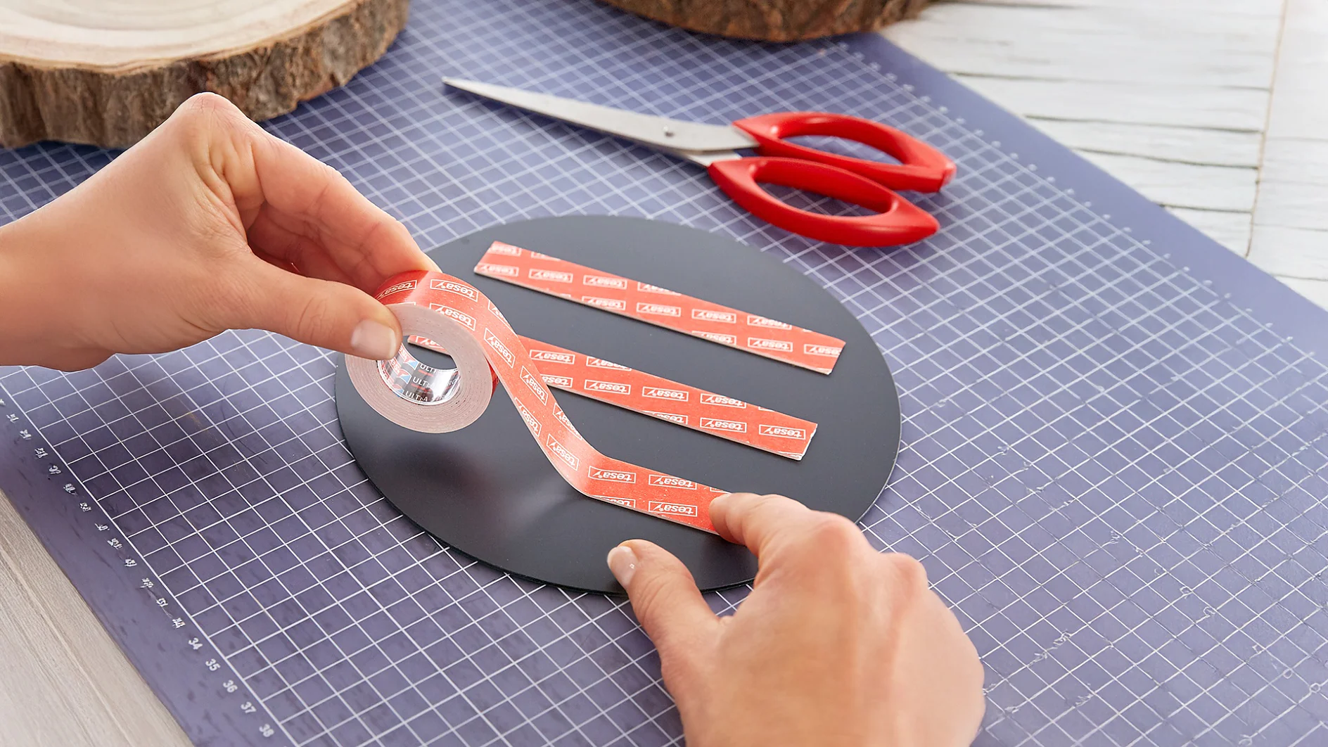 Een persoon brengt dubbelzijdig klevend tesa-tape aan op een rond zwart oppervlak op een snijmat, met een schaar met rood handvat en houten schijfjes op de achtergrond. (This text has been generated by AI)
