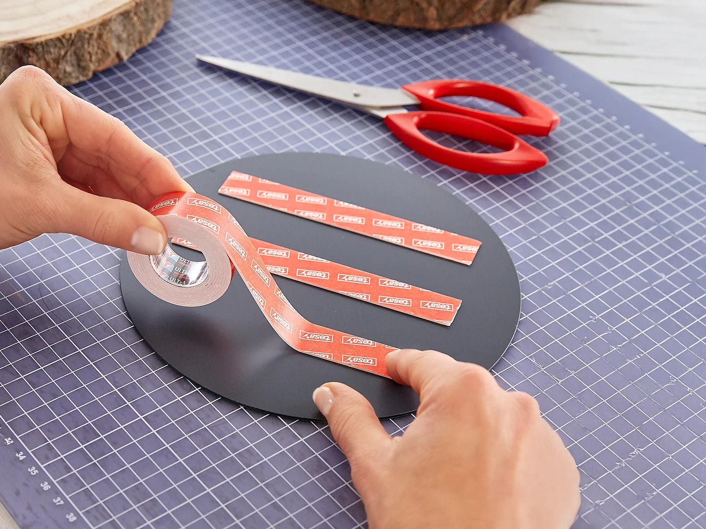 Een persoon brengt dubbelzijdig klevend tesa-tape aan op een rond zwart oppervlak op een snijmat, met een schaar met rood handvat en houten schijfjes op de achtergrond. (This text has been generated by AI)