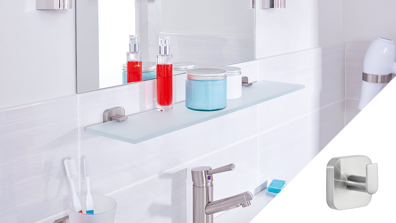 A modern bathroom counter with a glass shelf holding two bottles and a jar, one red and one blue. A mirror is above the shelf. Toothbrushes and a soap dispenser are on the counter. A small metallic wall hook, secured with tesa tape, is shown in a separate inset. (This text has been generated by AI)