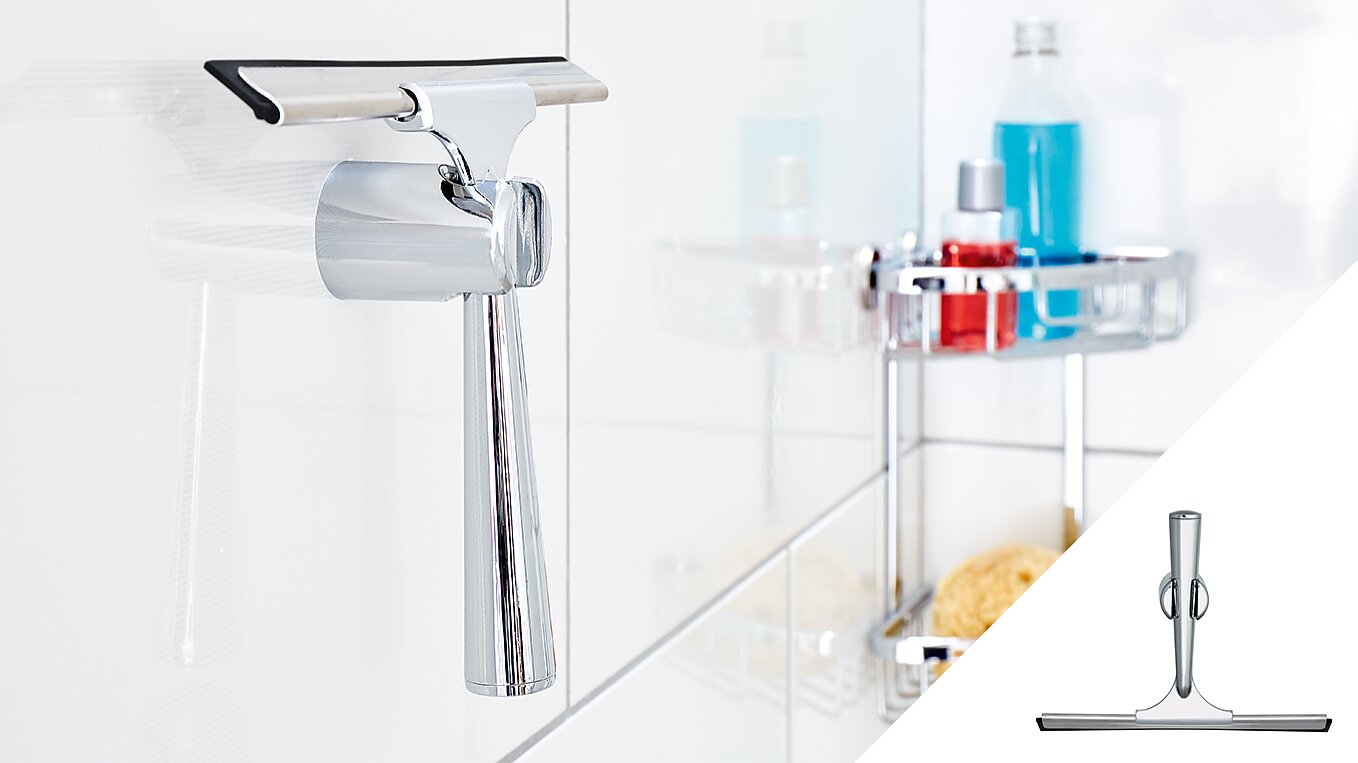 A metallic shower squeegee hangs on a hook attached to a tiled wall using tesa tape. In the background, a shower caddy holds various bottles and toiletries. An inset at the bottom right provides a close-up of the squeegee. (This text has been generated by AI)