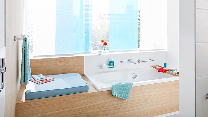 A modern bathroom features a wooden-accented bathtub next to a window with blue curtains. A bath tray holds a red book and blue towel. Toiletries and a potted plant are arranged neatly on the shelf. The space is bright and well-organized, with tesa tape ensuring seamless finishes throughout the room. (This text has been generated by AI)