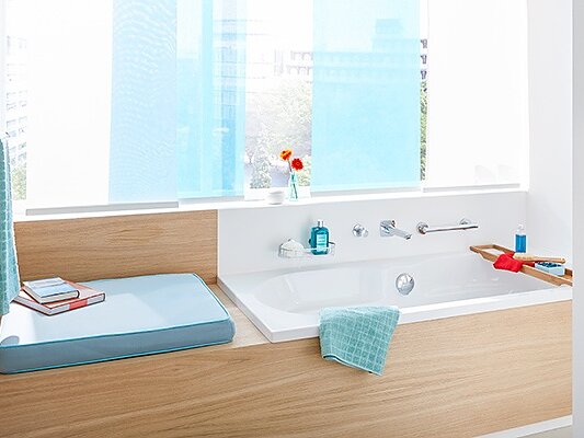 A modern bathroom features a wooden-accented bathtub next to a window with blue curtains. A bath tray holds a red book and blue towel. Toiletries and a potted plant are arranged neatly on the shelf. The space is bright and well-organized, with tesa tape ensuring seamless finishes throughout the room. (This text has been generated by AI)