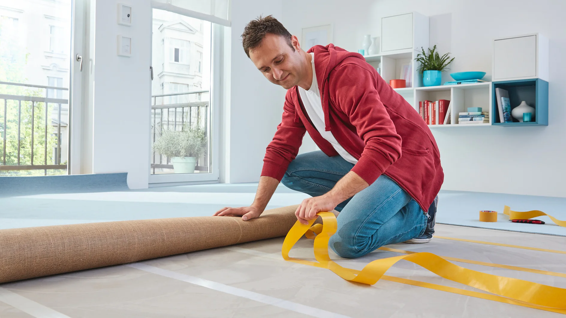 Een man met een rode capuchon knielt op de vloer en bereidt zich voor op het installeren van tapijt door het uit te rollen en gele dubbelzijdige tesa-tape aan te brengen op de vloer in een lichte, moderne kamer met planken en een groot raam. (This text has been generated by AI)