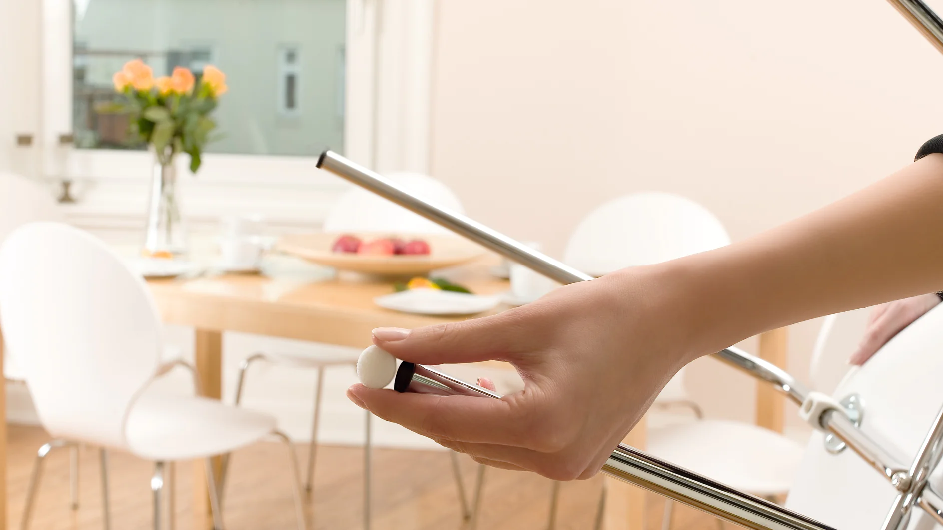 Een close-up van een hand die een metalen stoel in elkaar zet in een lichte eetkamer met een houten tafel, witte stoelen, een vaas met bloemen en op de achtergrond een fruitschaal. Hij zet de stoel in elkaar met behulp van tesa-tape. (This text has been generated by AI)