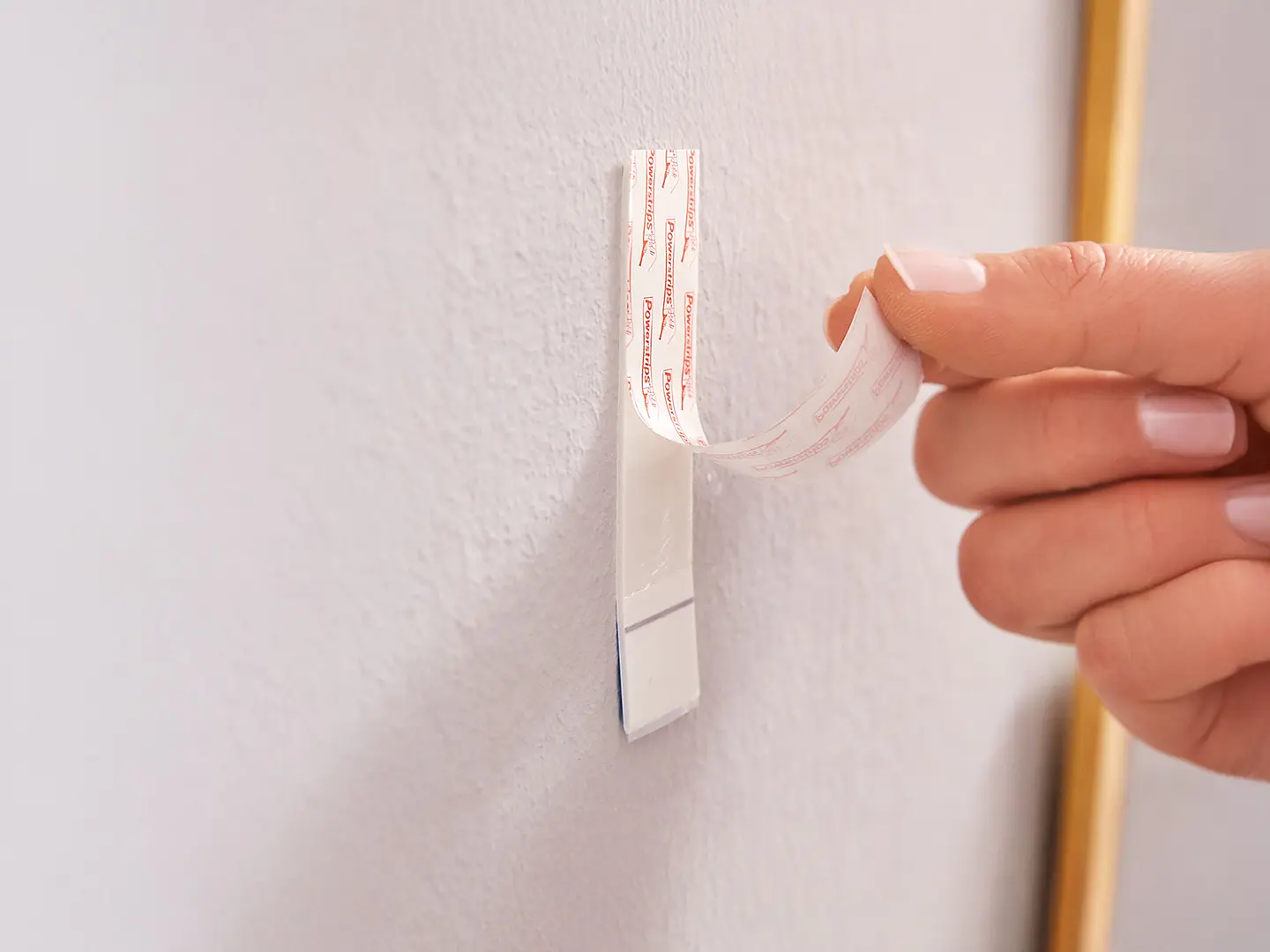 Een hand verwijdert de achterkant van een plakstrip van tesa tape op een witte haak die aan een lichtgekleurde muur is bevestigd, ter voorbereiding op het monteren of ophangen van iets.
