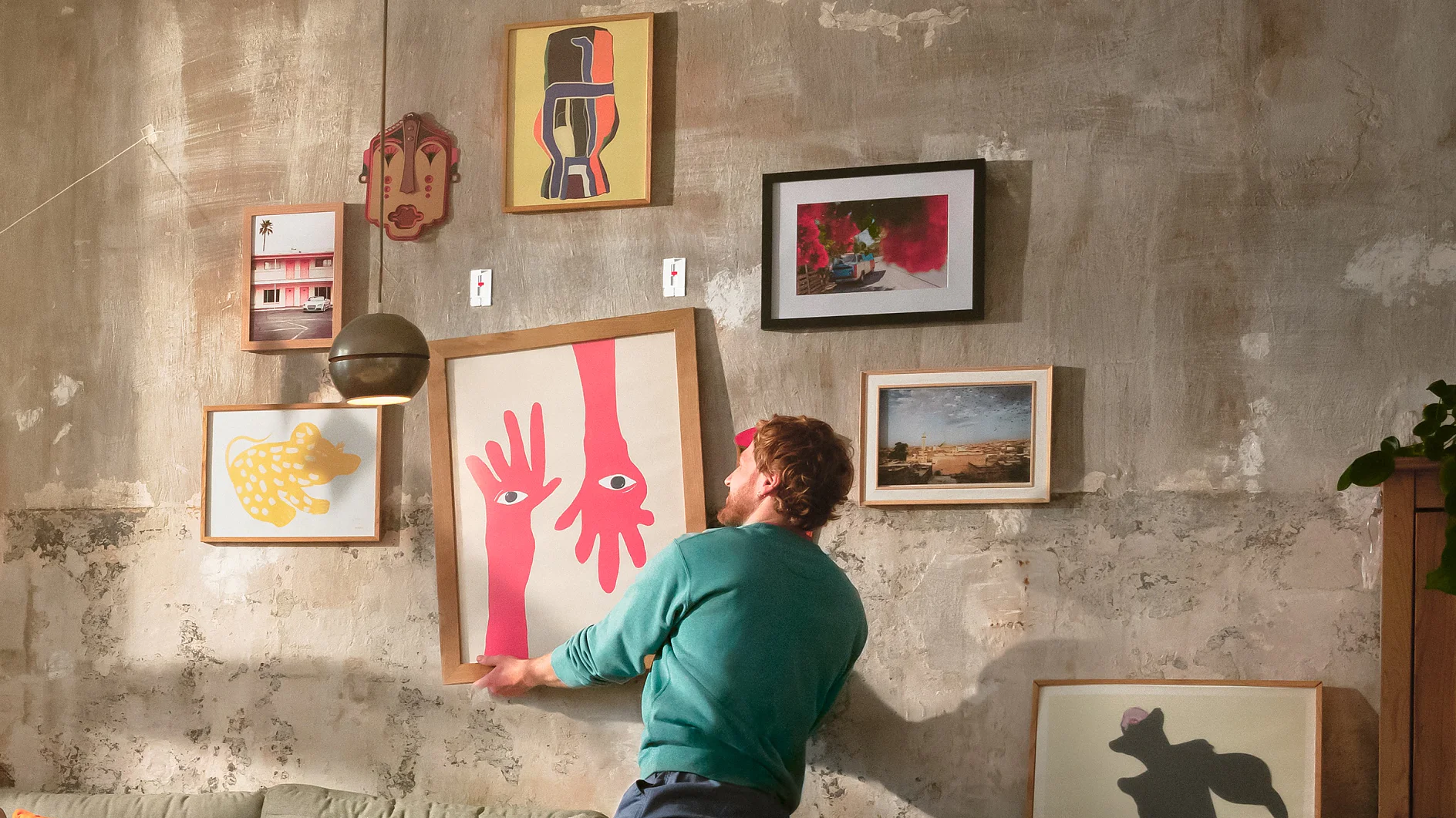 Una persona está de pie en un sofá, ajustando una pieza de arte enmarcada en una pared texturizada cubierta con varias imágenes y pinturas eclécticas en diferentes marcos, todos asegurados con cinta tesa. (This text has been generated by AI)