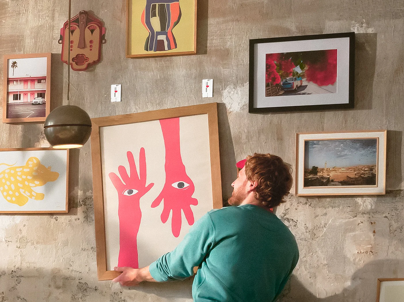 Una persona está de pie en un sofá, ajustando una pieza de arte enmarcada en una pared texturizada cubierta con varias imágenes y pinturas eclécticas en diferentes marcos, todos asegurados con cinta tesa. (This text has been generated by AI)