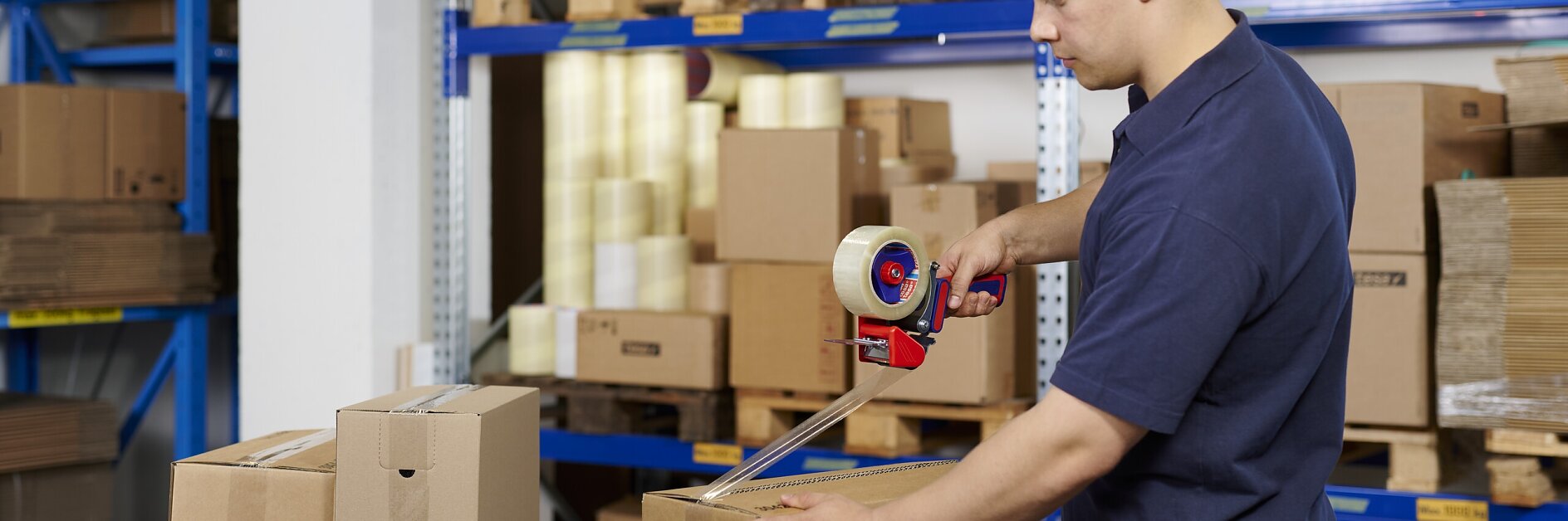 man in marineblauw uniform die kartonnen dozen dichtplakt met verpakkingstape in een magazijn met rekken vol gestapelde dozen en verpakkingsmaterialen