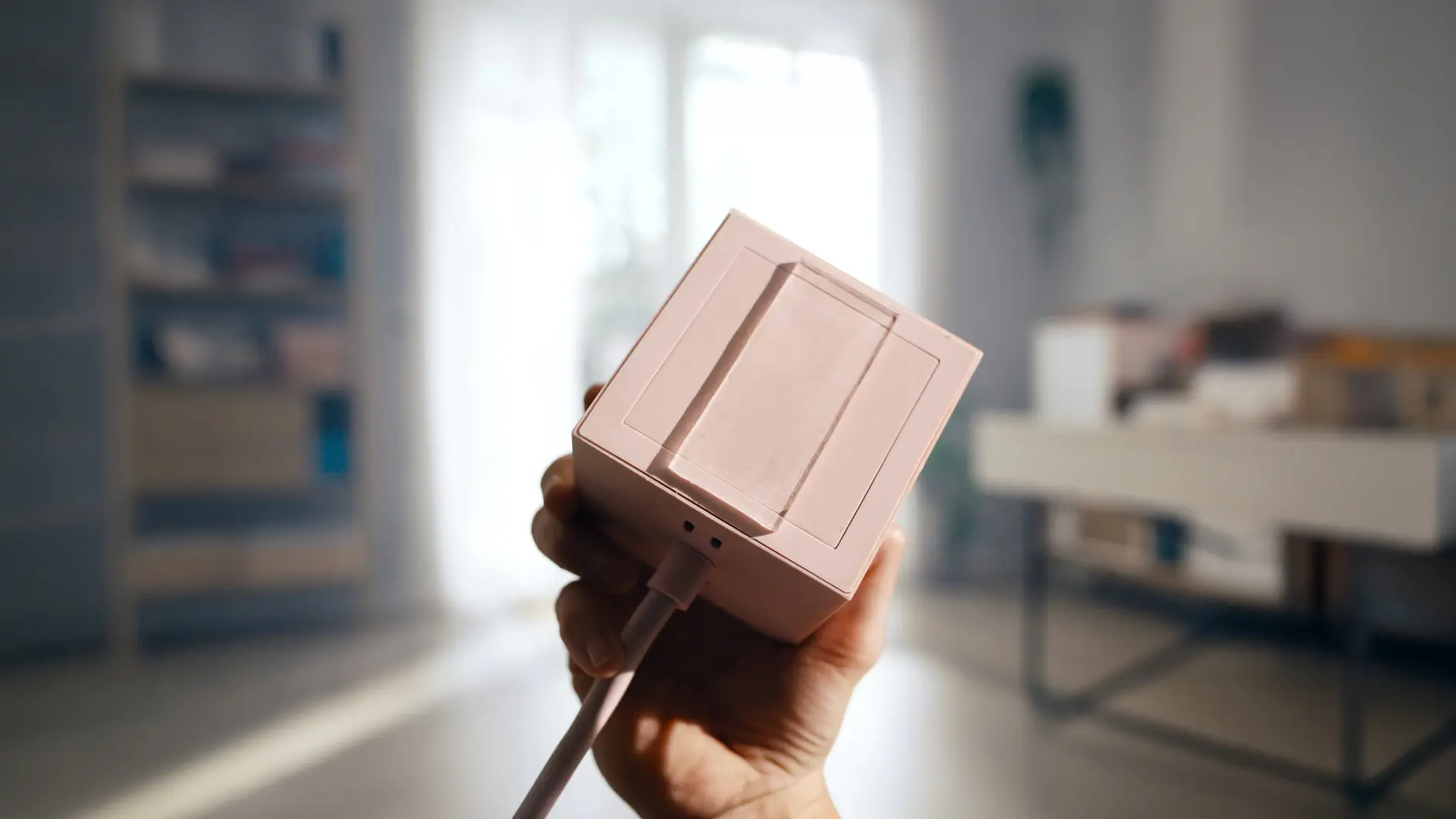 Eine Hand hält ein kleines, rosa, kastenförmiges elektronisches Gerät mit einem Kabel und tesa-Klebeband in einem hellen Raum mit unscharf abgebildeten Möbeln und einem Fenster.