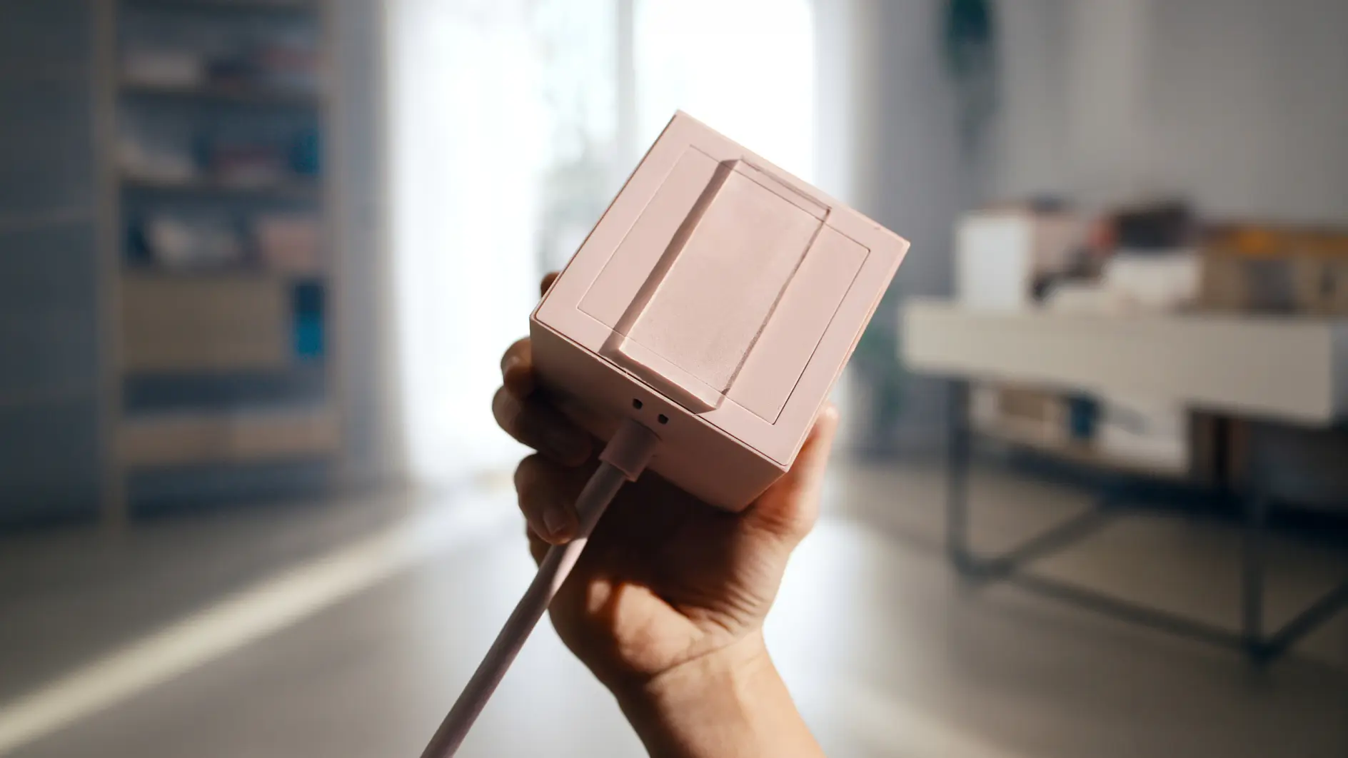 In einem gut beleuchteten Raum mit unscharf abgebildeten Regalen hält eine Hand ein kleines, rosa, rechteckiges elektronisches Gerät, an dem ein Kabel und ein Stück tesa-Klebeband befestigt sind. (This text has been generated by AI)