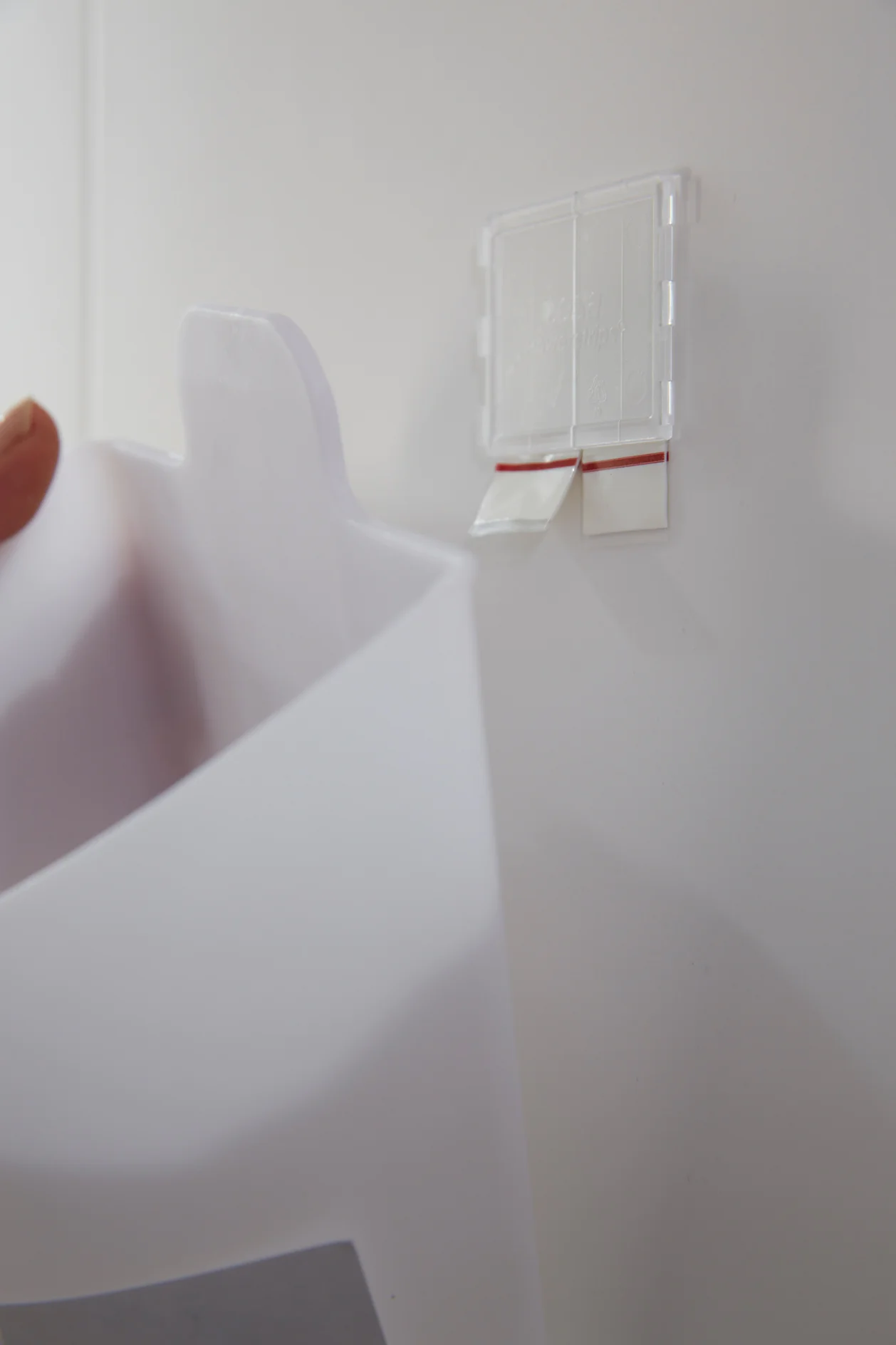 Biały haczyk Command Strip jest przymocowany do ściany. Ręka osoby wiesza na haczyku biały plastikowy uchwyt. Na haczyku widoczna jest czerwona taśma tesa. Ściana i uchwyt są jasne. (This text has been generated by AI)