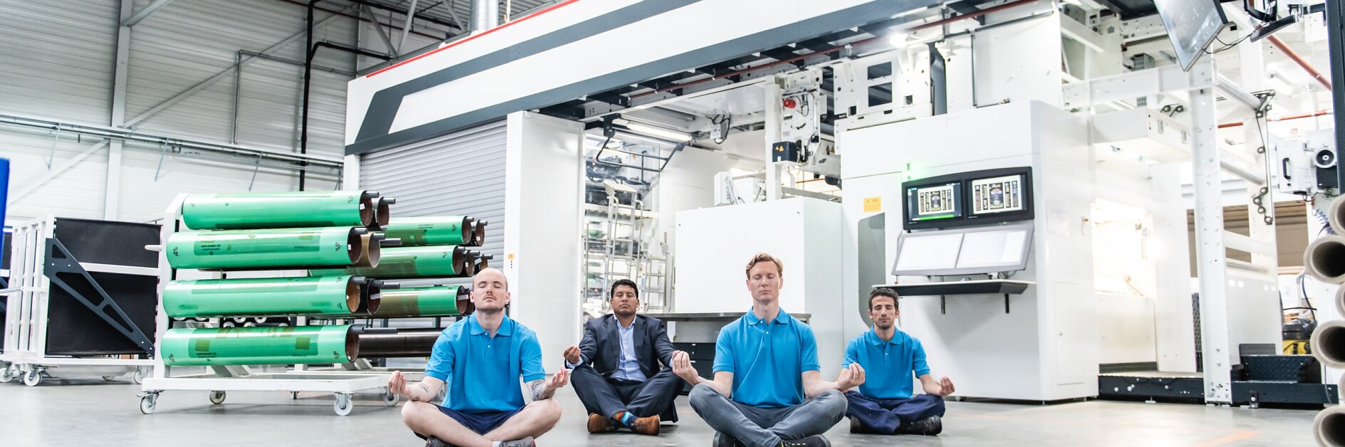 Four people sit cross-legged and meditate on the floor of an industrial workshop. They are wearing blue shirts and are positioned in front of large machinery and rolls of tesa tape material. The setting is well-lit with industrial equipment visible.