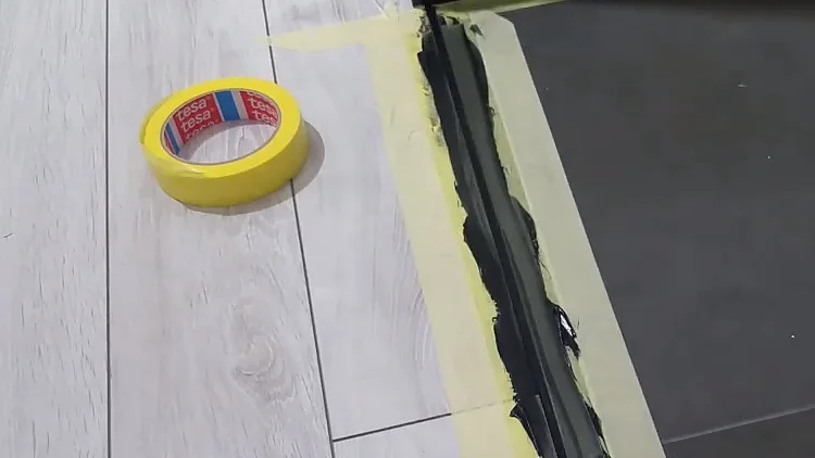 Rolka żółtej taśmy tesa jest umieszczona na jasnej drewnianej podłodze obok czarnej pomalowanej szczeliny. Żółta taśma tesa jest przyklejona wzdłuż krawędzi pomalowanego obszaru, który graniczy z ciemnymi płytkami. (This text has been generated by AI)