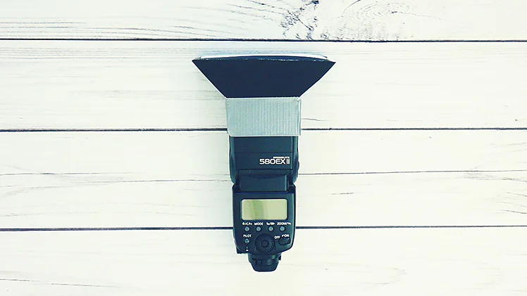 Lampa błyskowa aparatu fotograficznego z przymocowanym czarnym i srebrnym dyfuzorem jest umieszczona na białej drewnianej powierzchni. Cyfrowy wyświetlacz i elementy sterujące są widoczne z przodu urządzenia, przymocowane na miejscu za pomocą taśmy tesa. (This text has been generated by AI)