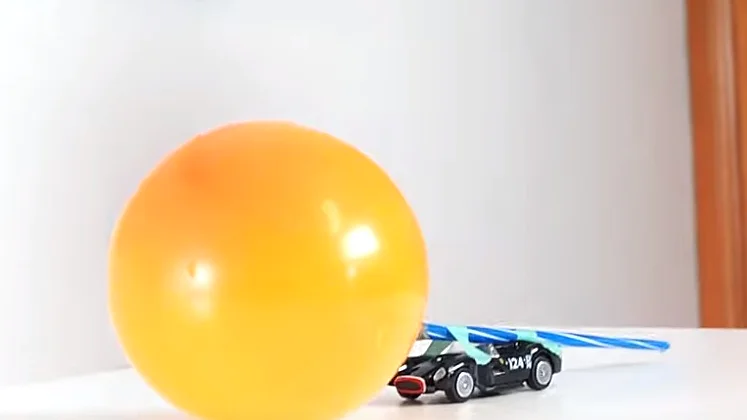 Na stole stoi mały czarny samochodzik z niebieskimi słomkami. Za samochodzikiem nadmuchany jest pomarańczowy balon, co sugeruje domowy system napędowy z użyciem taśmy tesa. Tło to zwykłe wnętrze. (This text has been generated by AI)