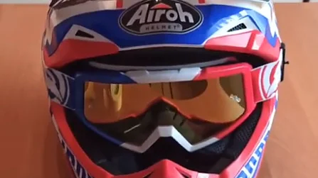 Na stole położono kask motocrossowy z żywym czerwonym, niebieskim i białym wzorem. W części twarzowej ma parę pomarańczowych gogli, a na górze zamontowano kamerę akcji za pomocą taśmy tesa. (This text has been generated by AI)