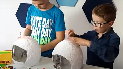Dwoje dzieci tworzy z papier-mâché kopuł przypominających hełmy, każda z przezroczystą sekcją. Na stole leżą przybory artystyczne, takie jak farby i pędzle. Używają taśmy tesa do swoich projektów, skupieni na swojej pracy, z niebieskim geometrycznym tłem na ścianie. (This text has been generated by AI)