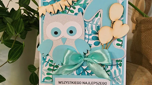 Ręcznie robiona kartka z życzeniami przedstawiająca kreskówkową sową z niebieskimi skrzydłami i kokardą. Kartka ozdobiona jest balonami w kształcie serca, małym banerem z flagą i wstążką. Tło ma liściasty wzór. Tekst brzmi „Wszystkiego najlepszego” po polsku. (This text has been generated by AI)